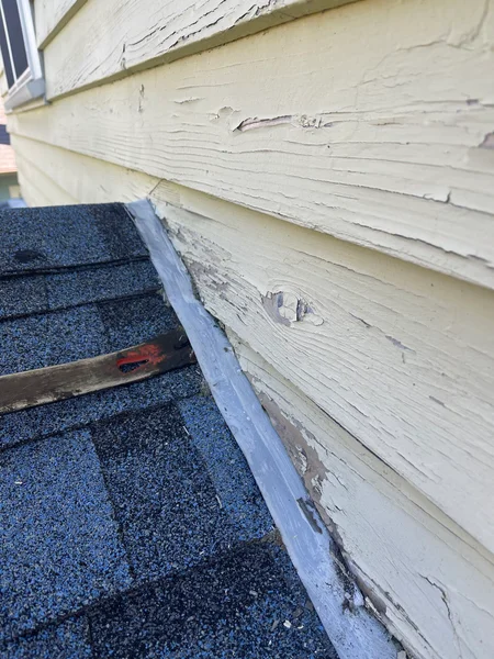 Damaged wood siding with peeling paint next to roofing shingles showing deterioration and potential dry rot